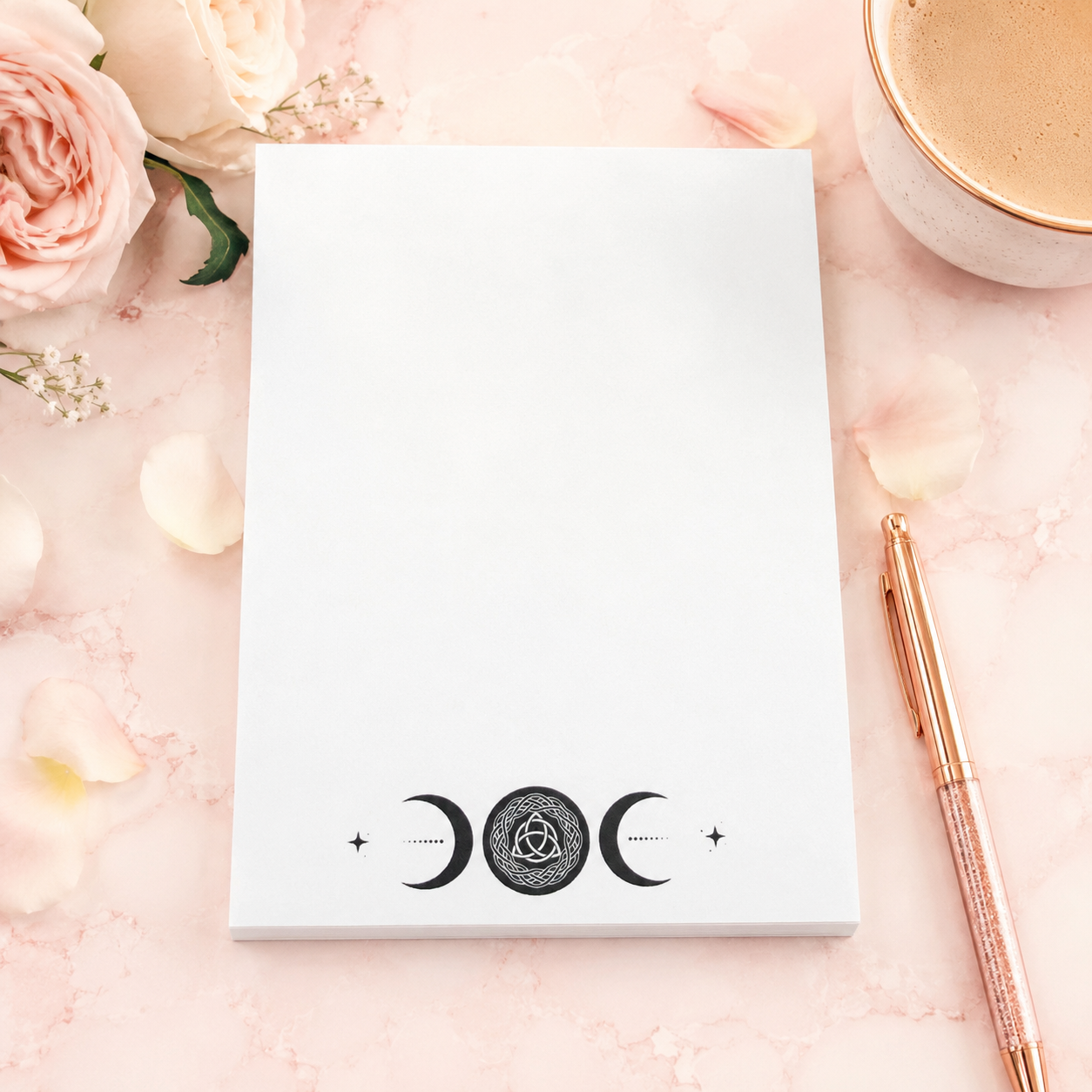 White triple moon notepad with Celtic knot moon symbol and celestial moon phase design on a soft pink feminine desktop with rose gold pen and floral accents.