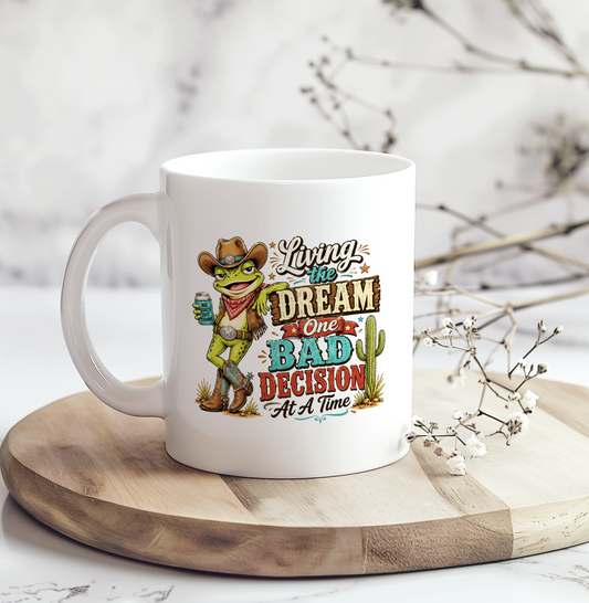 White ceramic coffee mug featuring a funny cowboy frog holding a drink with the phrase "Living the Dream One Bad Decision At A Time," perfect as a funny western coffee mug gift for frog lovers and country humor fans.