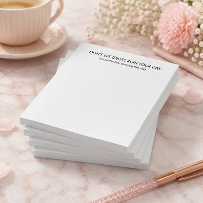 Funny notepad with the phrase "Don't Let Idiots Ruin Your Day" displayed on a blush pink desktop with floral accents, coffee cup, and rose gold pen.