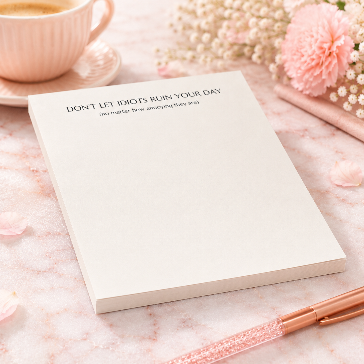 Funny notepad with the phrase "Don't Let Idiots Ruin Your Day" displayed on a blush pink desktop with floral accents, coffee cup, and rose gold pen.