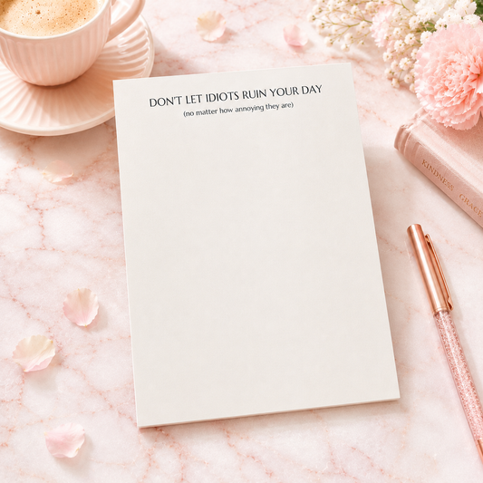 Funny notepad with the phrase "Don't Let Idiots Ruin Your Day" displayed on a blush pink desktop with floral accents, coffee cup, and rose gold pen.