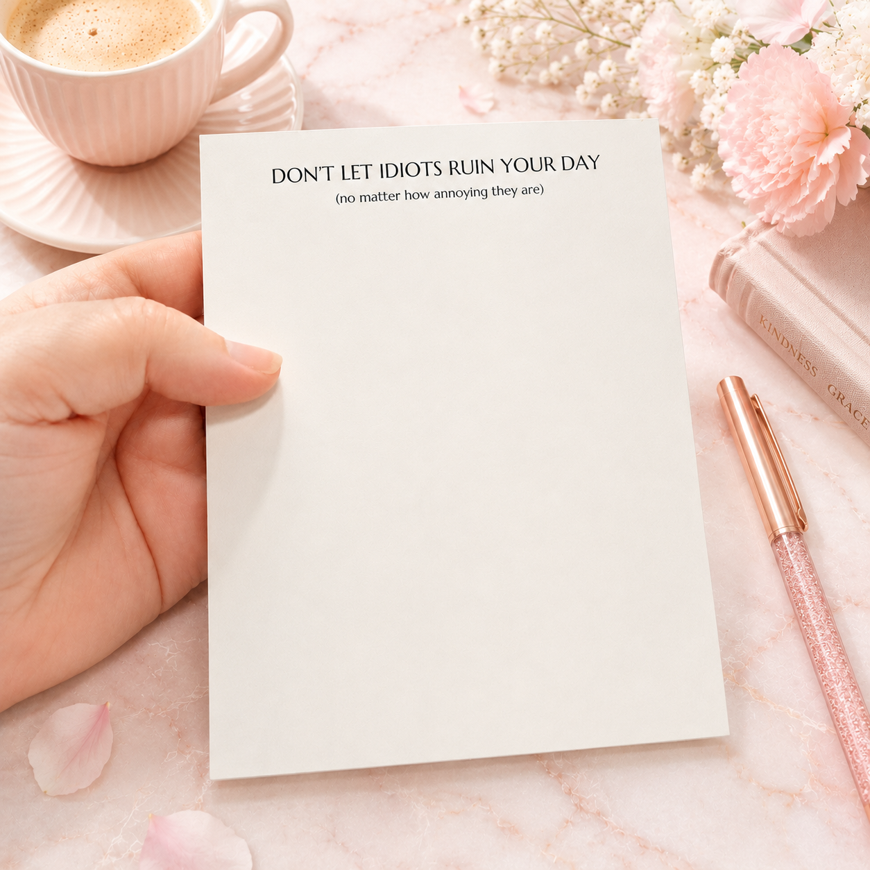 Funny notepad with the phrase "Don't Let Idiots Ruin Your Day" displayed on a blush pink desktop with floral accents, coffee cup, and rose gold pen.