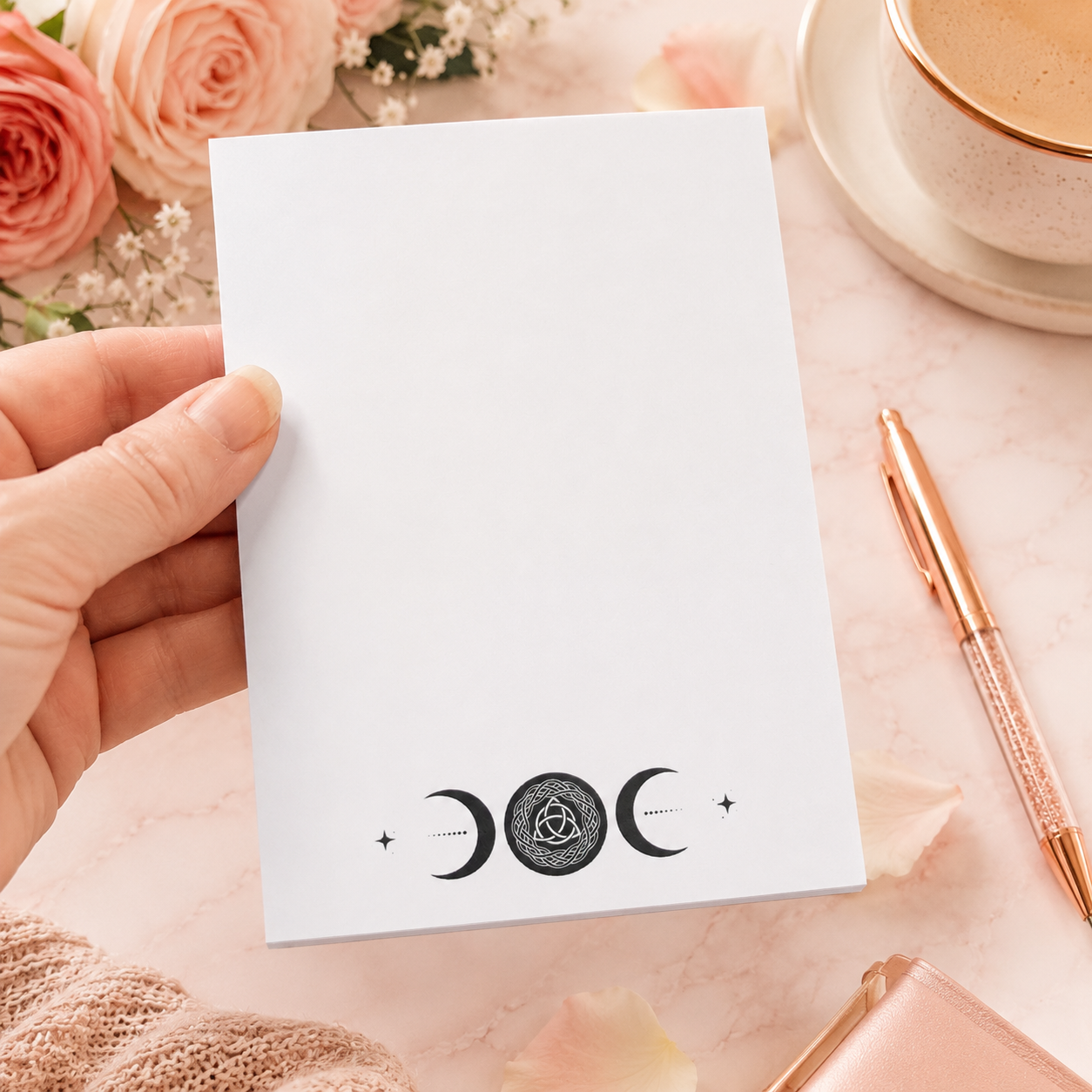 White triple moon notepad with Celtic knot moon symbol and celestial moon phase design on a soft pink feminine desktop with rose gold pen and floral accents.